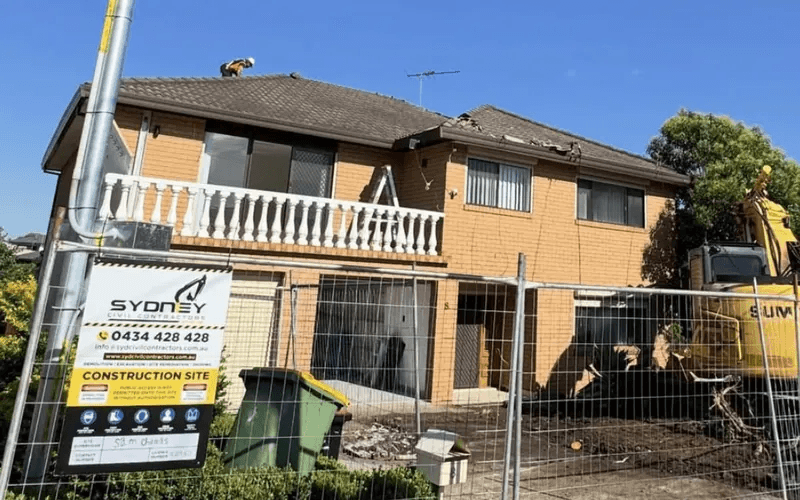 A Sydney Civil Contractors site sign on temporary fencing in front of a two storey brick house mid demolition