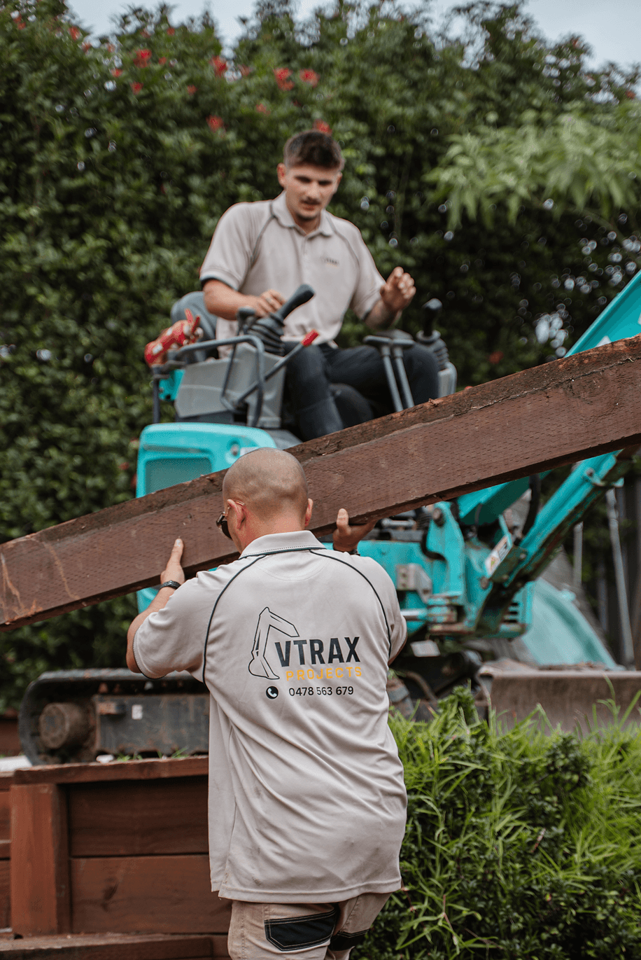 A VTRAX Projects crew member in a branded shirt lifting a timber sleeper on a backyard retaining wall job